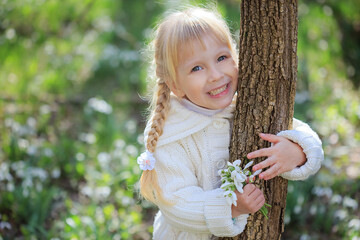 Beautiful little girl with a bouquet of snowdrops. A little girl in a white knitted sweater hugs a tree trunk. Bright sunny spring day