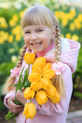 Cute little girl holding a bouquet of yellow tulips on the background of beautiful flowers. A girl in a pink coat.