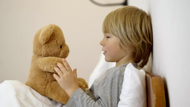 Side view of carefree Caucasian boy lying in bed talking with teddy bear. Portrait of cute relaxed child sharing secrets with toy at bedtime at home. Childhood and leisure.