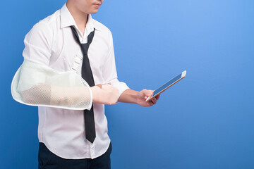 Young businessman with an injured arm in a sling using a tablet over blue background in studio, insurance and healthcare concept