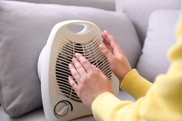 Woman warming hands near fan heater indoors, closeup