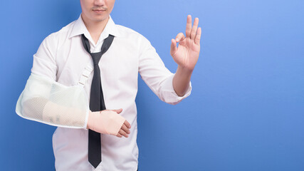 Young businessman with an injured arm in a sling over blue background in studio, insurance and...