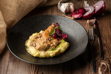 Baked meat and mashed potatoes served with mushrooms, green sauce and lettuce in black bowl. Close-up on a wooden background with a sacking, sliced onion, cloves of garlic and silver cutlery by side.
