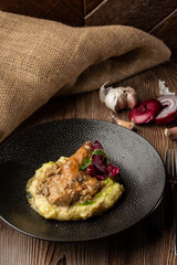 Baked meat and mashed potatoes served with mushrooms, green sauce and lettuce in black bowl. Close-up on a wooden background with a sacking, sliced onion, cloves of garlic and silver cutlery by side.