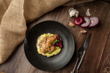 Baked meat and mashed potatoes served with mushrooms, green sauce and lettuce in black bowl. Close-up on a wooden background with a sacking, sliced onion, garlic and silver cutlery by side. Top view.