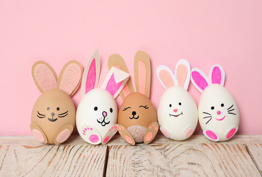 Eggs as cute bunnies on white wooden table against pink background. Easter celebration