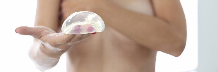 Nude female patient holds silicone breast implant in her hand. Plastic surgery mammaplasty breast correction concept.
