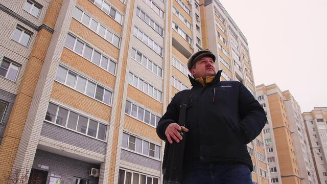 Caucasian man with a bag on his shoulder waiting for a taxi in the yard. Male in a warm jacket with a fur hat outside in cool weather. In the background, multi-storey high-rise buildings.