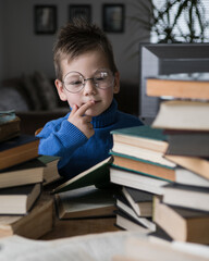 Five year old boy in glasses reading a book with a stack of books next to him. Smart intelligent preschool kid choosing books to borrow.