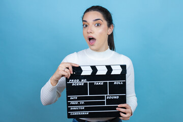 Young caucasian woman wearing white sweater over blue background holding clapperboard very happy...