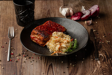 Baked meat served with red sauce, fermented cabbage and dill in black bowl. Close-up on a wooden background with a black glass, sliced onion, cloves of garlic, mix of pepper seeds and cutlery by side.