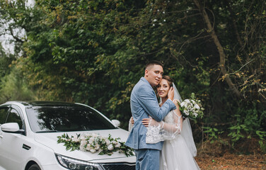 A stylish, tall groom in a blue striped suit hugs a cute curly-haired bride in a white lace dress, standing on an asphalt road against the backdrop of a chic, expensive, decorated car.
