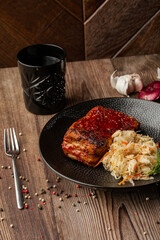 Baked meat served with red sauce, sauerkraut and dill in black bowl. Close-up on wooden background with a black glass, sliced onion, cloves of garlic, mix of pepper seeds and a fork by side.