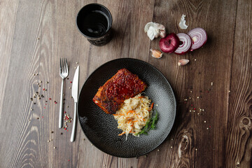 Baked meat served with red sauce, sauerkraut and dill in black bowl. Close-up on a wooden background with a black glass, onion, garlic, mix of pepper seeds and cutlery by side. Top view, copy space.