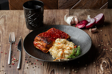 Baked meat served with red sauce, fermented cabbage and dill in black bowl. Close-up on a wooden background with a black glass, sliced onion, cloves of garlic, mix of pepper seeds and cutlery by side.