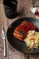 Baked meat served with red sauce, sauerkraut and dill in black bowl. Close-up on wooden background with a black glass, sliced onion, cloves of garlic, mix of pepper seeds and a fork by side.