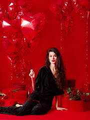 Valentine's Day. Beauty girl in black with red heart-shaped balloons having fun, on a red background