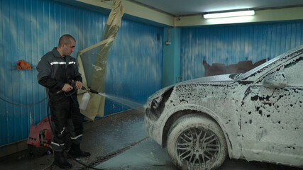 A Caucasian man works in a car wash service. A man in a worker's uniform sprays a car with detergent