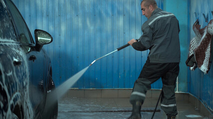 A Caucasian man works in a car wash service. A man in a worker's uniform washes the car thoroughly by spraying it with a water pistol.