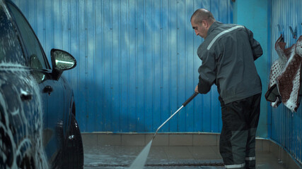 A Caucasian man works in a car wash service. A man in a worker's uniform washes the car thoroughly by spraying it with a water pistol.