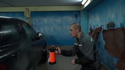 A Caucasian man works in a car wash service. A man in a worker's uniform washes the car thoroughly by spraying it with a water pistol.