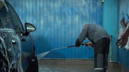 A Caucasian man works in a car wash service. A man in a worker's uniform washes the car thoroughly by spraying it with a water pistol.