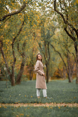 A girl with a Euro-Asian appearance in a pink coat and blue jeans with holes walks in an autumn park