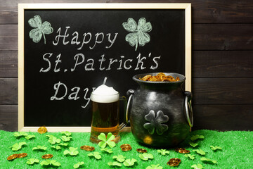 Happy St. Patricks Day chalk lettering on blackboard, mug of beer, leprechauns pot of gold treasure and shamrock leaves and coins on green grass, dark wooden planks background. Saint Patricks Day