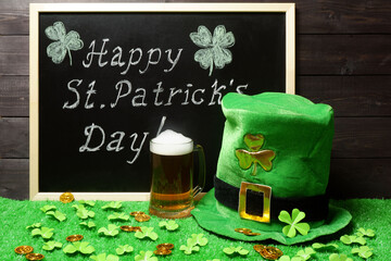 Happy St. Patricks Day chalk lettering on blackboard, mug of beer, leprechauns hat and shamrock leaves and coins on green grass, dark wooden planks background. Saint Patricks Day template