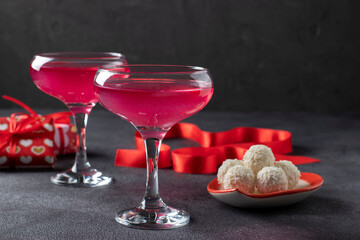 Two glasses of shimmering pink champagne, coconut candies in a heart-shaped plate and gifts against a dark background. Romantic dinner on Valentine's Day.