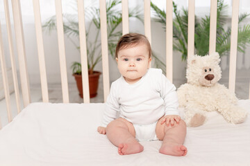 baby six months sitting in a crib in a white bodysuit with toys Teddy bears