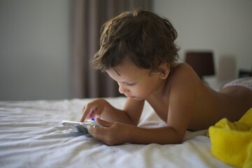 A small child lies on the bed and looks at the phone.