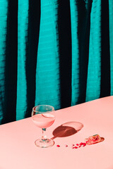 Single glass of wine with creative drink and pills look a like or sweer candy lay on a floor with small bottle against pink background and blue cyan silk fabric curtain. Modern direct  light