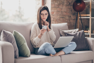 Photo of minded serious young woman hold hand chin read phone laptop sit couch indoors in house...