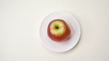 Red apple on the plate. Beautiful colorful fruit of the apple-tree on a white dish. The daily healthy diet not only for the nutrition of vegetarians or vegans. A top view on the white background.
