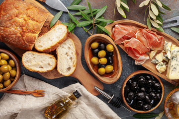 Black and green Olives, olive tree branch, oil, cheese Dorblu or Gorgonzola, prosciutto and ciabatta bread on dark  table. Top view. Flat lay