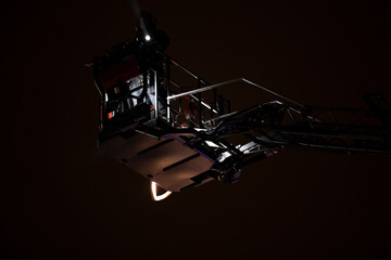 Firefighters rise on a mechanical sliding ladder to the epicenter of the fire. Fighting fire from bucket atop a fire truck. A fireman's crane in action, silhouette against a dark night sky.