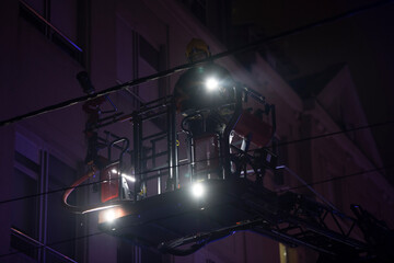 Firefighters rise on a mechanical sliding ladder to the epicenter of the fire. Fighting fire from bucket atop a fire truck. A fireman's crane in action, silhouette against a dark night sky.