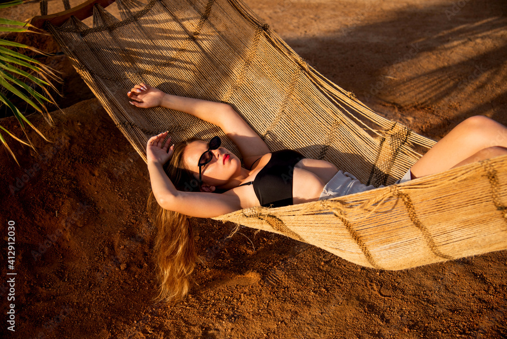 Wall mural young beautiful girl in sunglasses posing while lying on a hammock, beautiful woman resting on vacat