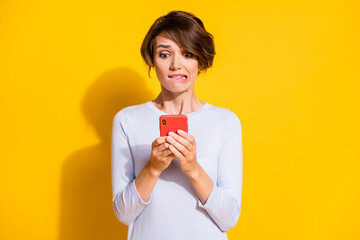 Photo portrait of scared girl biting lower lip holding phone in two hands isolated on bright yellow colored background