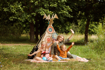 Mom and her daughters waving hands sitting next to wigwam decoration in the park