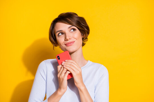 Photo portrait of dreamy girl looking at blank space holding phone in two hands isolated on bright yellow colored background