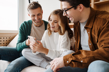 Happy gay family smiling and using mobile phone while sitting on couch