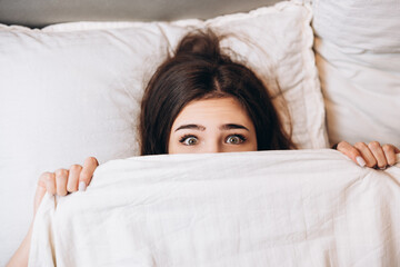 Obraz premium model woman hiding under the blanket. Young woman is very scared. Woman with big open eyes in bed