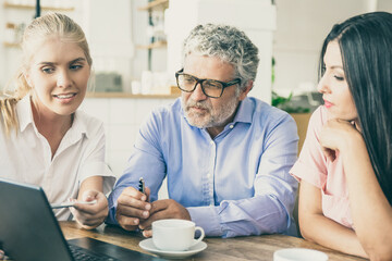 Positive female agent showing project presentation on laptop to young woman and mature man, pointing pen at display, explaining details. Consulting or communication concept