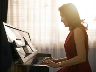 Middle age dignified Asian woman learning and studying new skills how to play the piano via online class from a tablet computer with happiness. Beautiful orange sunlight comes from window in the back
