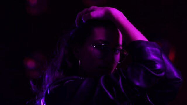 Beautiful brunette girl in club atmosphere posing cheerfully to the camera. Attractive young woman in sunglasses looking straight to the camera while standing on the pink neon and violet light.