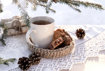 Vintage rustic card with white porcelain cup of tea and cake on white wooden table decorated with snowy fir tree and cones and lacy white napkin. Winter seasonal holiday background. Food and drink