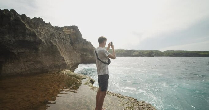 Circling handheld view of a male tourist at Broken Beach Pasih Uug in Bali, taking pictures of the view with his phone