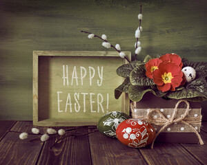Text Happy Easter. Zero waste Easter Easter arrangement in red, green and earth colors. Painted wooden eggs and spring flowers. Box with red primrose and pussy willow on rustic wood table..